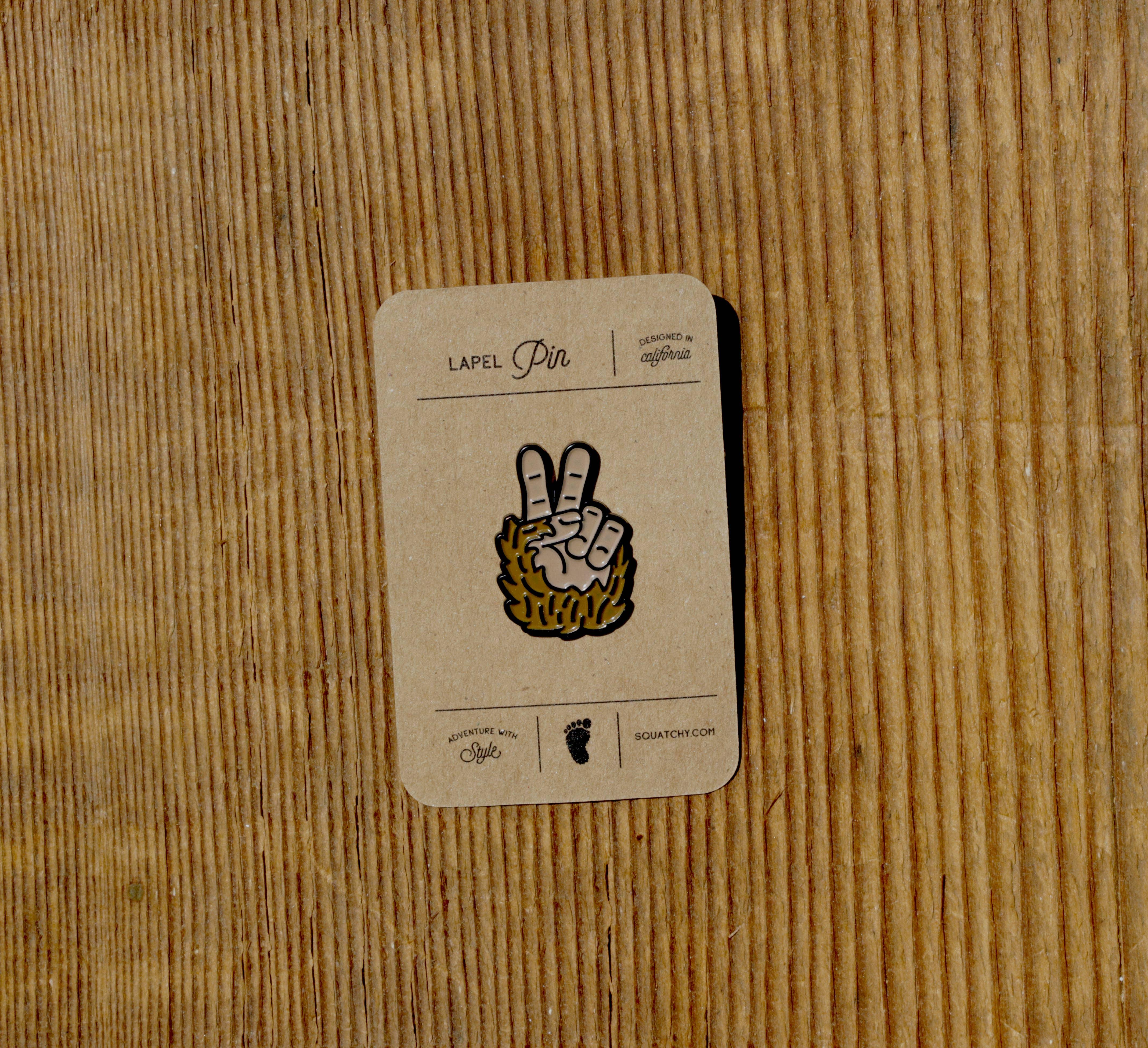 Squatchy lapel pin with hairy hand making peace sign, designed in California, on wooden surface