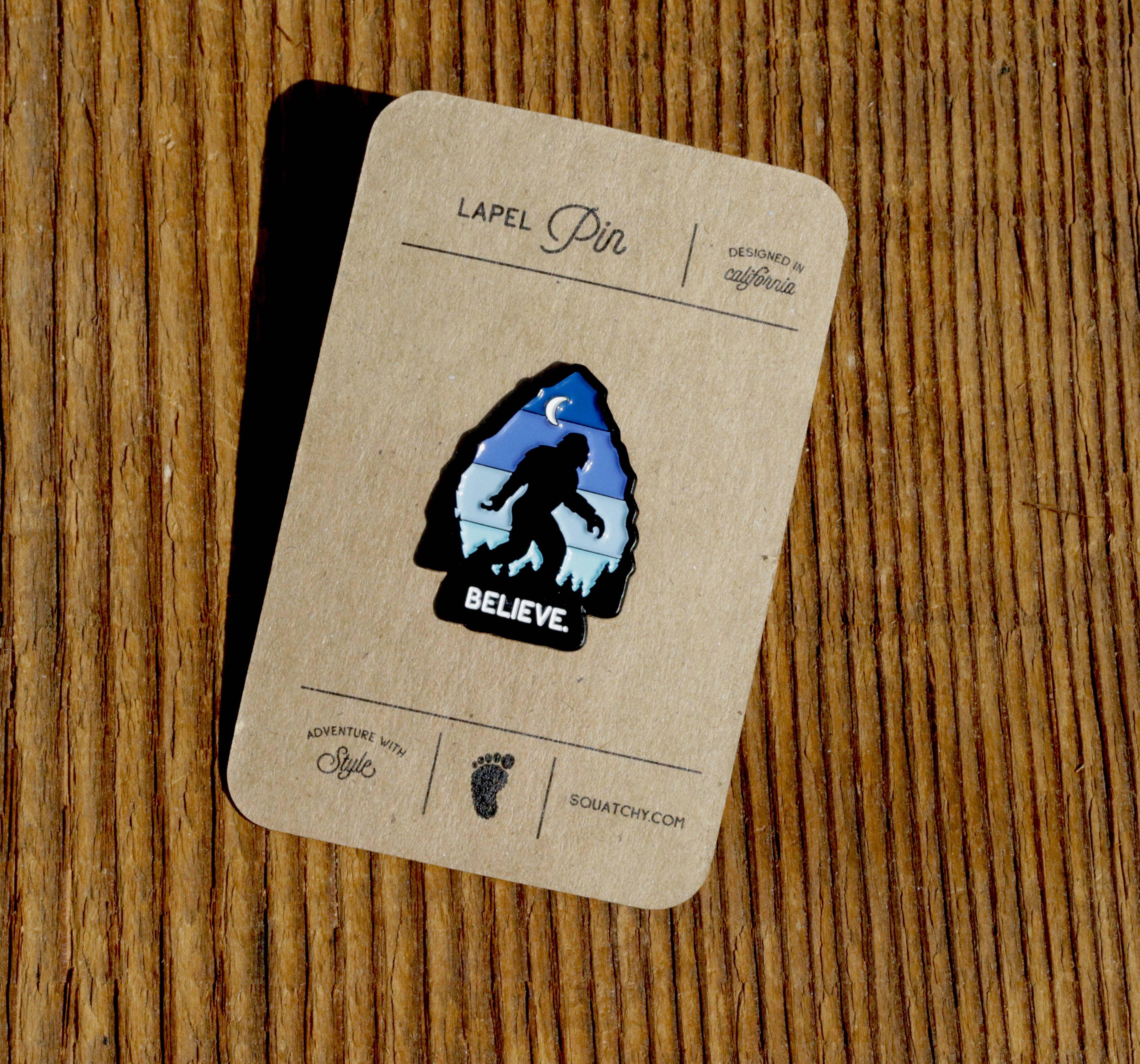 Sasquatch lapel pin with 'BELIEVE' text, designed in California, on wooden background