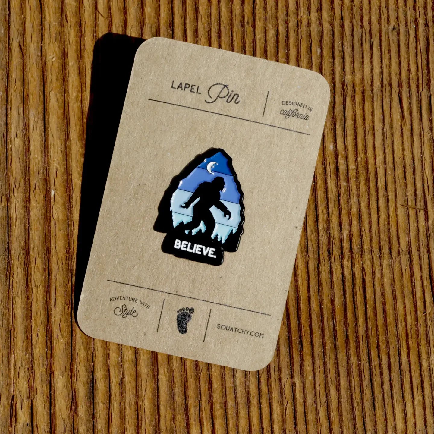 Sasquatch lapel pin with 'BELIEVE' text, designed in California, on wooden background