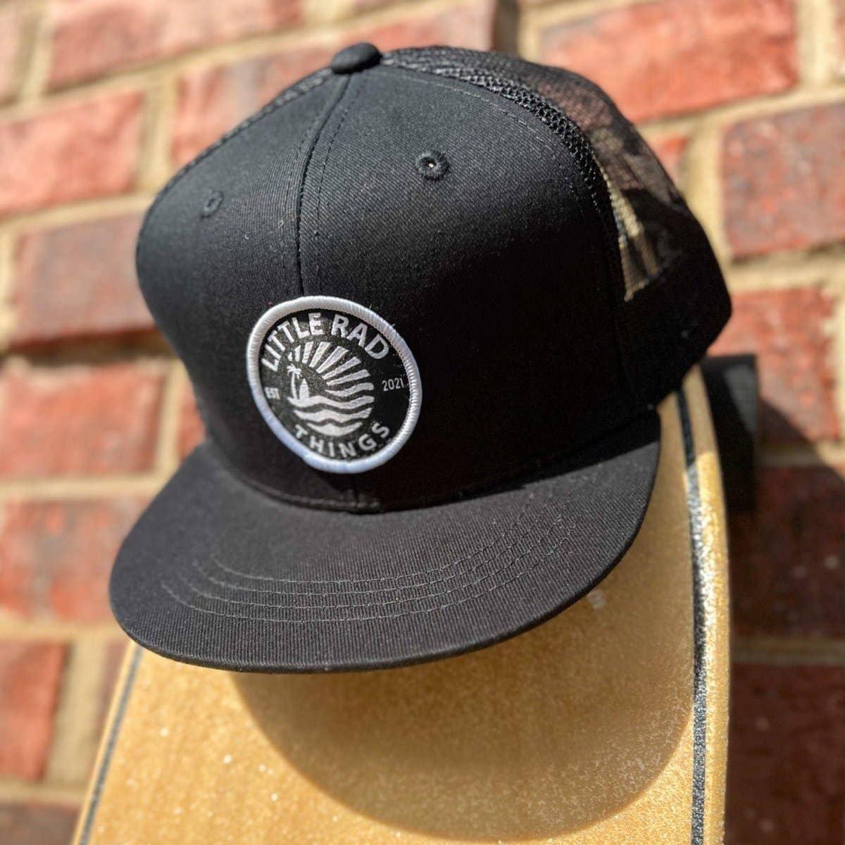 Black trucker hat with 'Little Rad Things' patch, resting on skateboard against brick wall