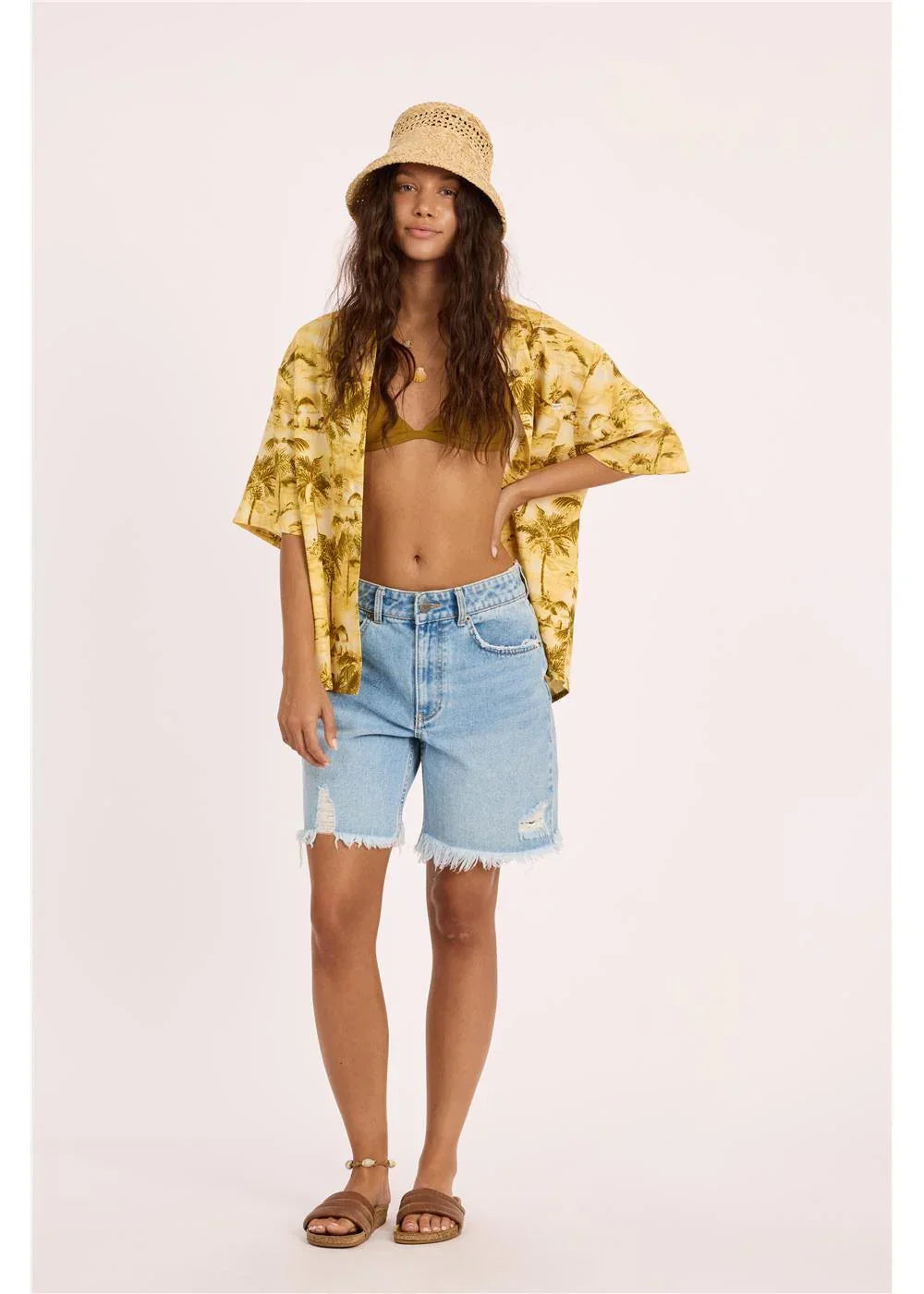 Woman in yellow palm print cover-up, denim shorts, straw hat, and sandals for beach or coastal style