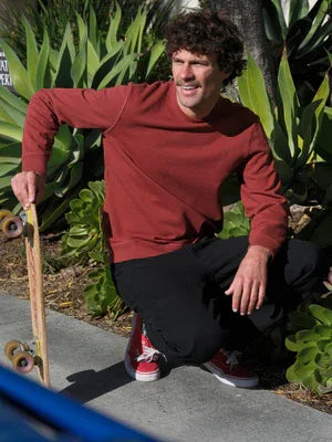 Man in red long-sleeve shirt crouching with skateboard outdoors near greenery, coastal PNW style