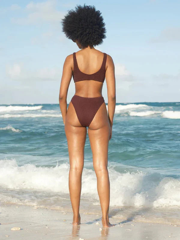 Woman in brown high-waisted bikini standing on beach facing ocean waves, coastal lifestyle swimwear