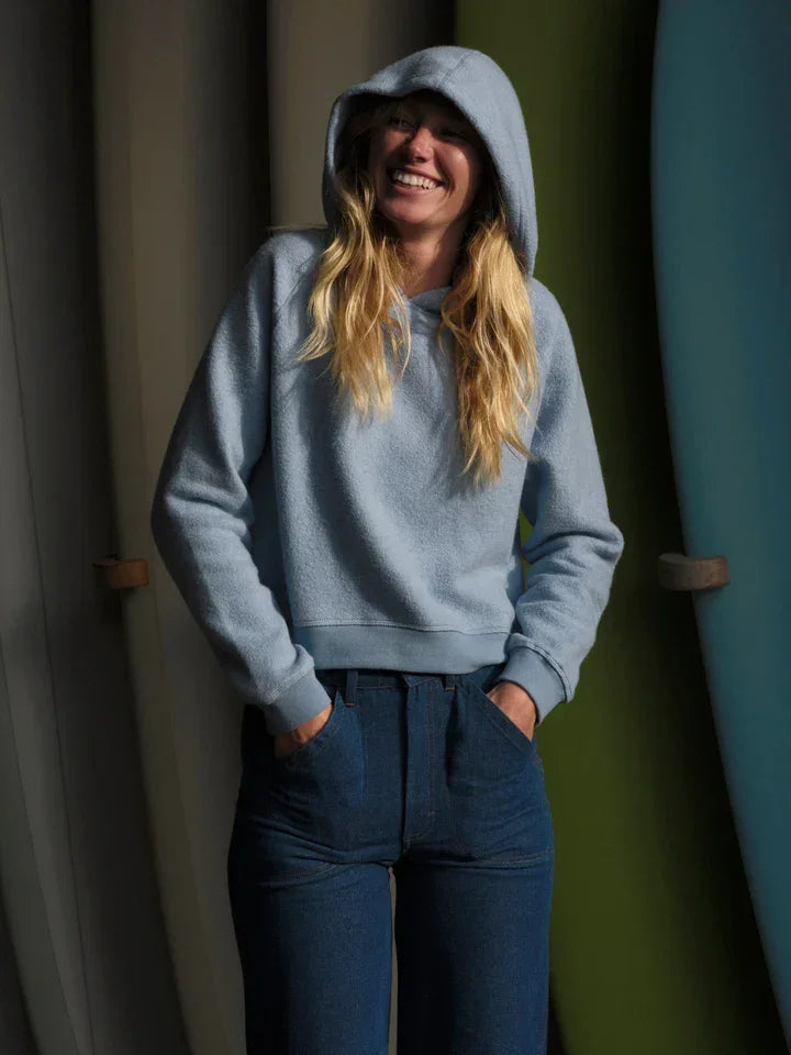 Woman in light blue hoodie and jeans, smiling with hands in pockets near surfboard