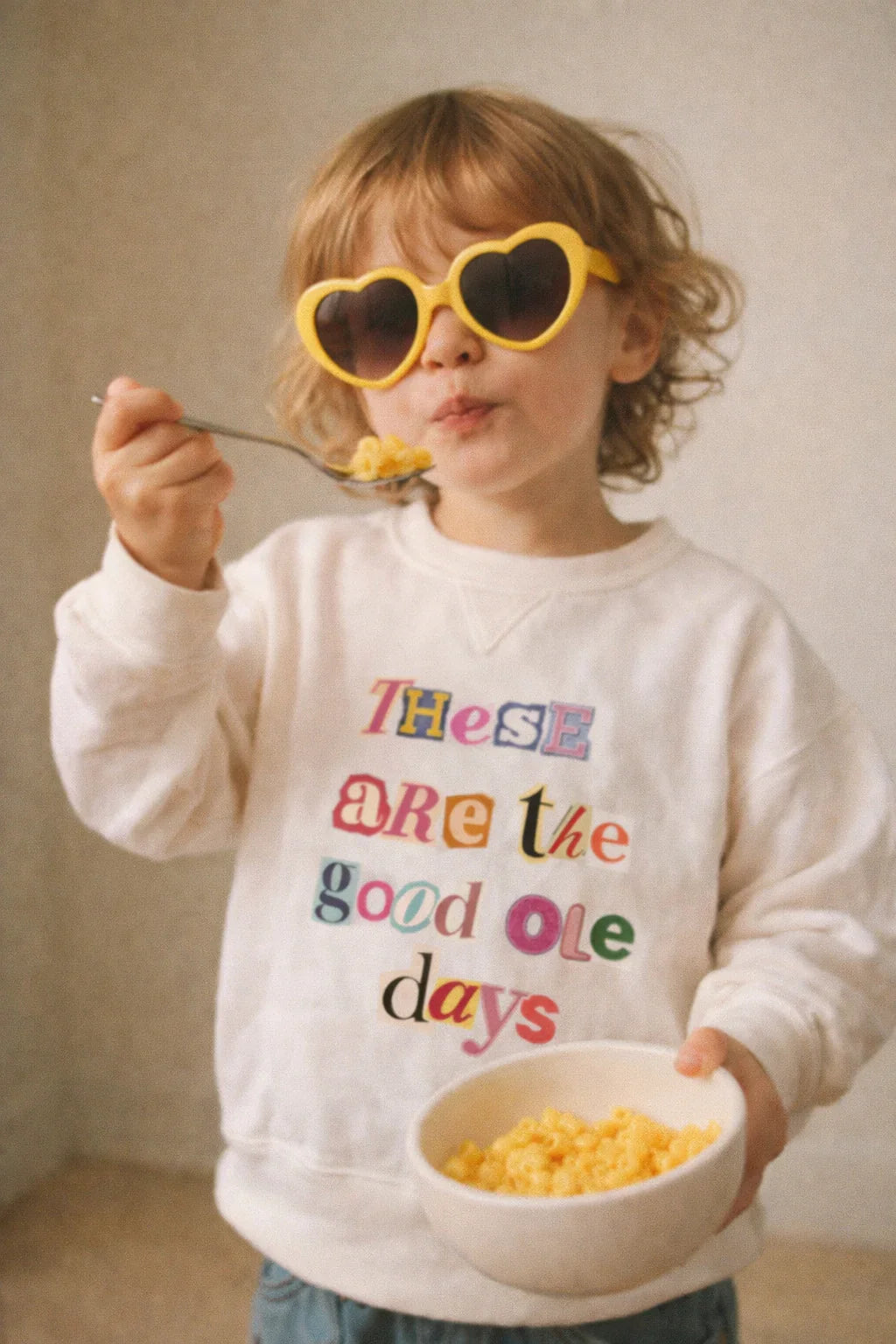 Toddler in 'These are the good ole days' sweatshirt eating mac and cheese, wearing heart-shaped sunglasses