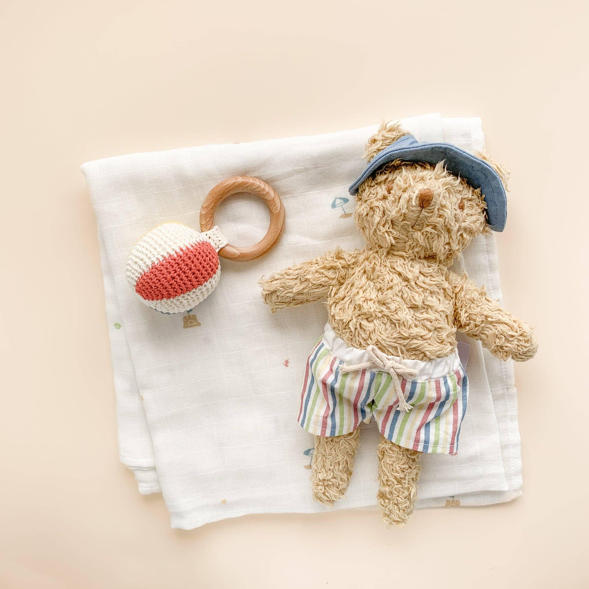 Cozy teddy bear in striped shorts and blue hat with knit hot air balloon rattle on muslin blanket