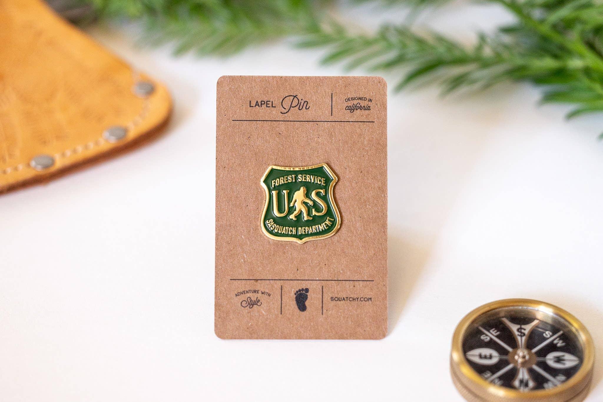 Forest Service Sasquatch Department lapel pin with green and gold badge, compass, and leather pouch