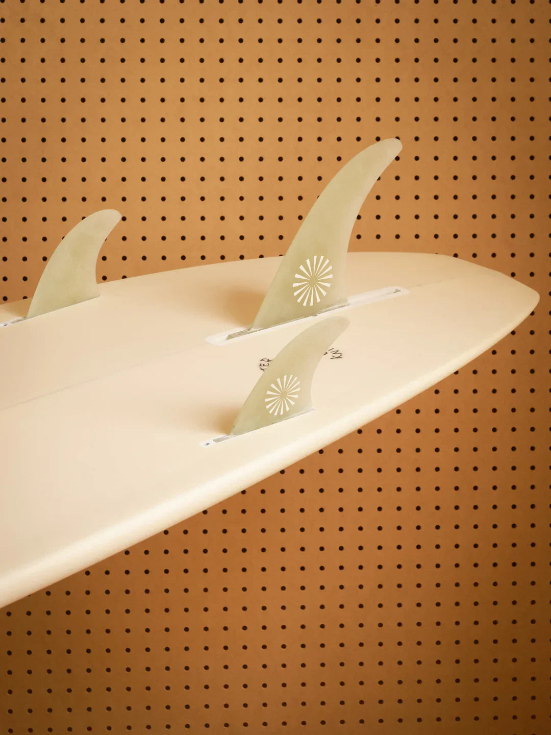 Cream surfboard with three fins featuring sunburst designs, displayed against a perforated brown wall.