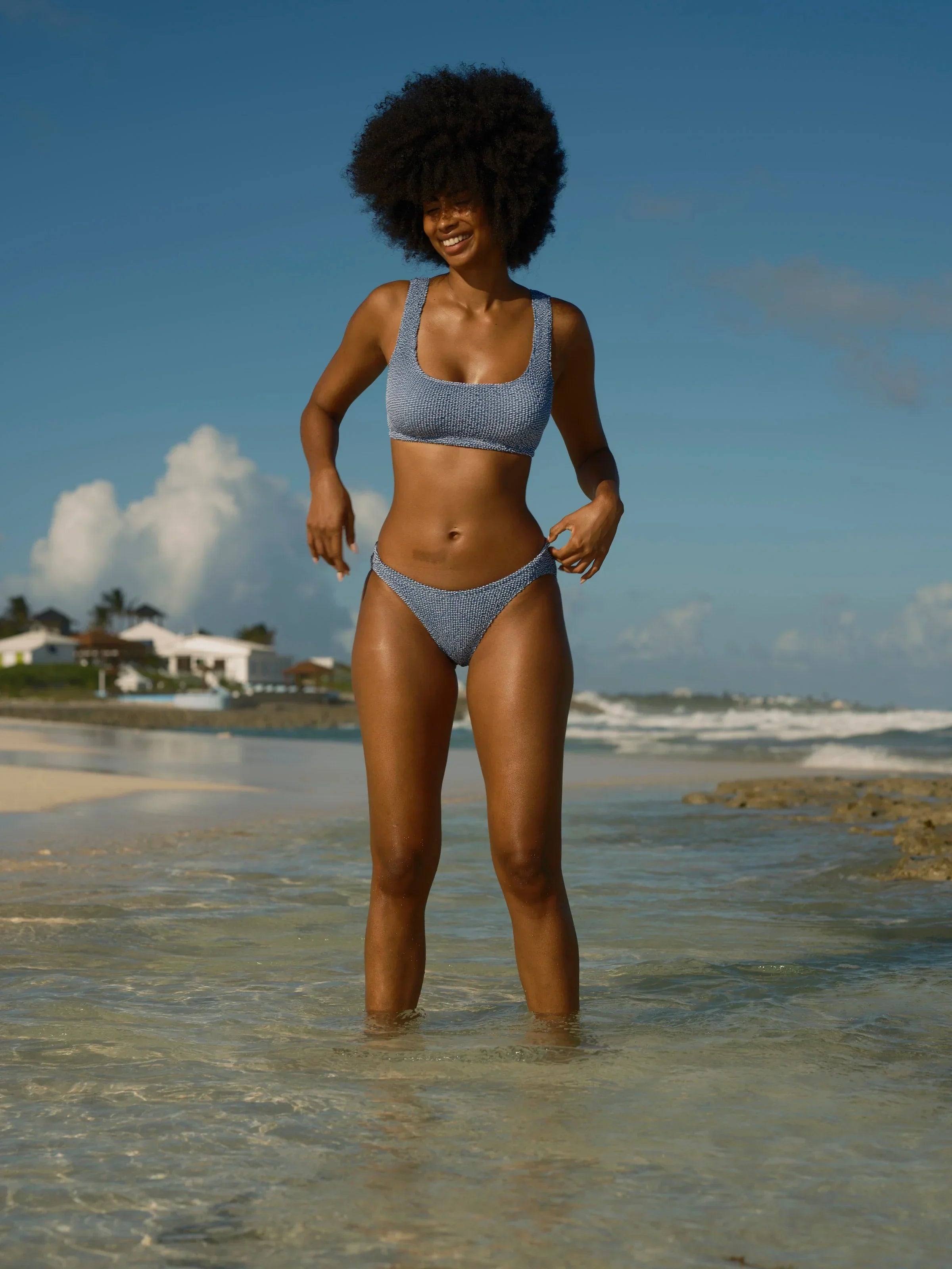 Woman in blue patterned bikini smiling on sunny beach, ocean waves behind, coastal lifestyle
