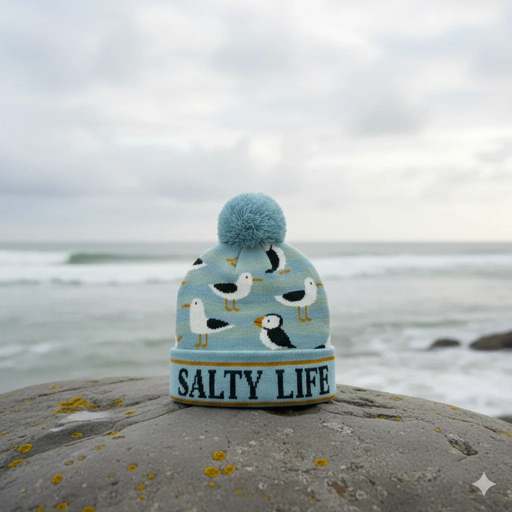 Light blue beanie with seagulls and 'SALTY LIFE' text, perched on coastal rock near ocean waves