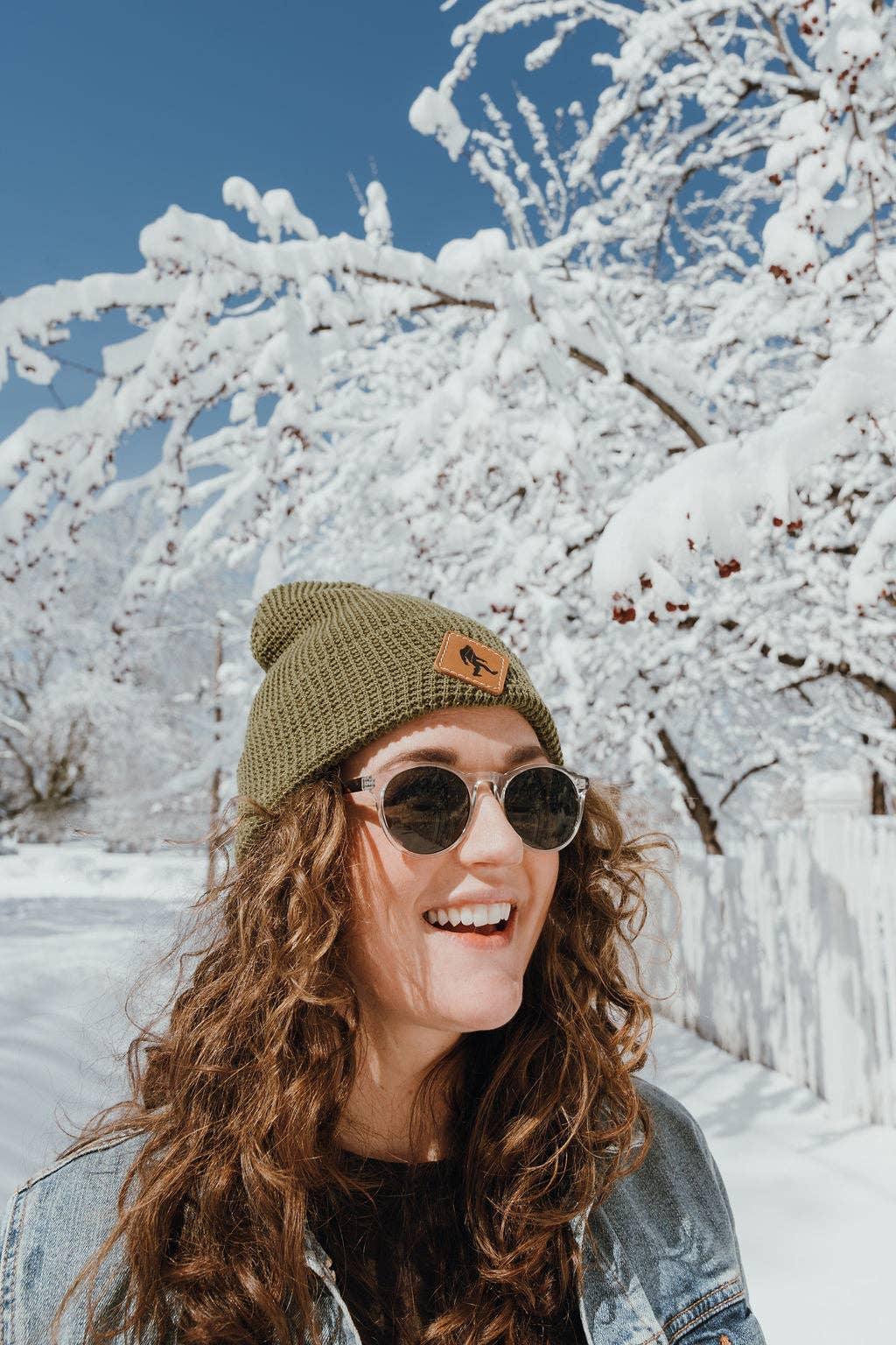 Woman in olive beanie and sunglasses smiles in snowy winter scene with snow-covered trees