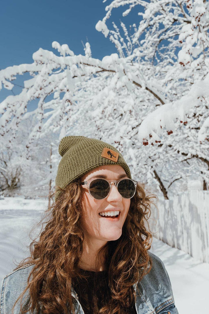 Woman in olive beanie and sunglasses smiles in snowy winter scene with snow-covered trees