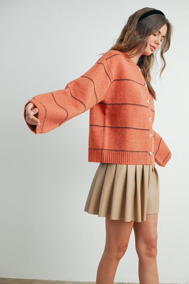 Woman in orange knit cardigan with contrast stitching, paired with beige pleated skirt, coastal style outfit