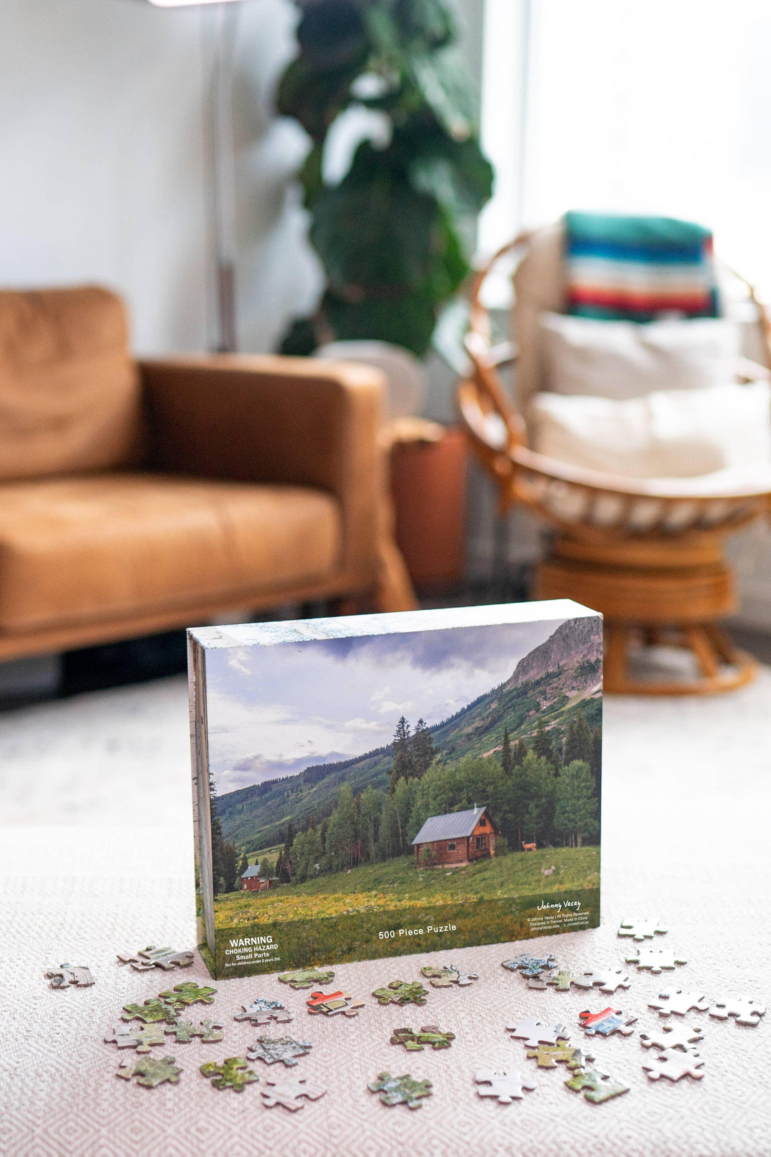 500-piece mountain cabin puzzle box with scattered pieces on rug, cozy living room backdrop