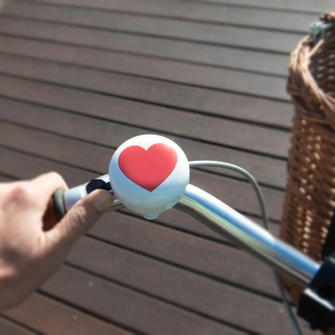 White bike bell with red heart on handlebar, wicker basket beside, wooden deck background