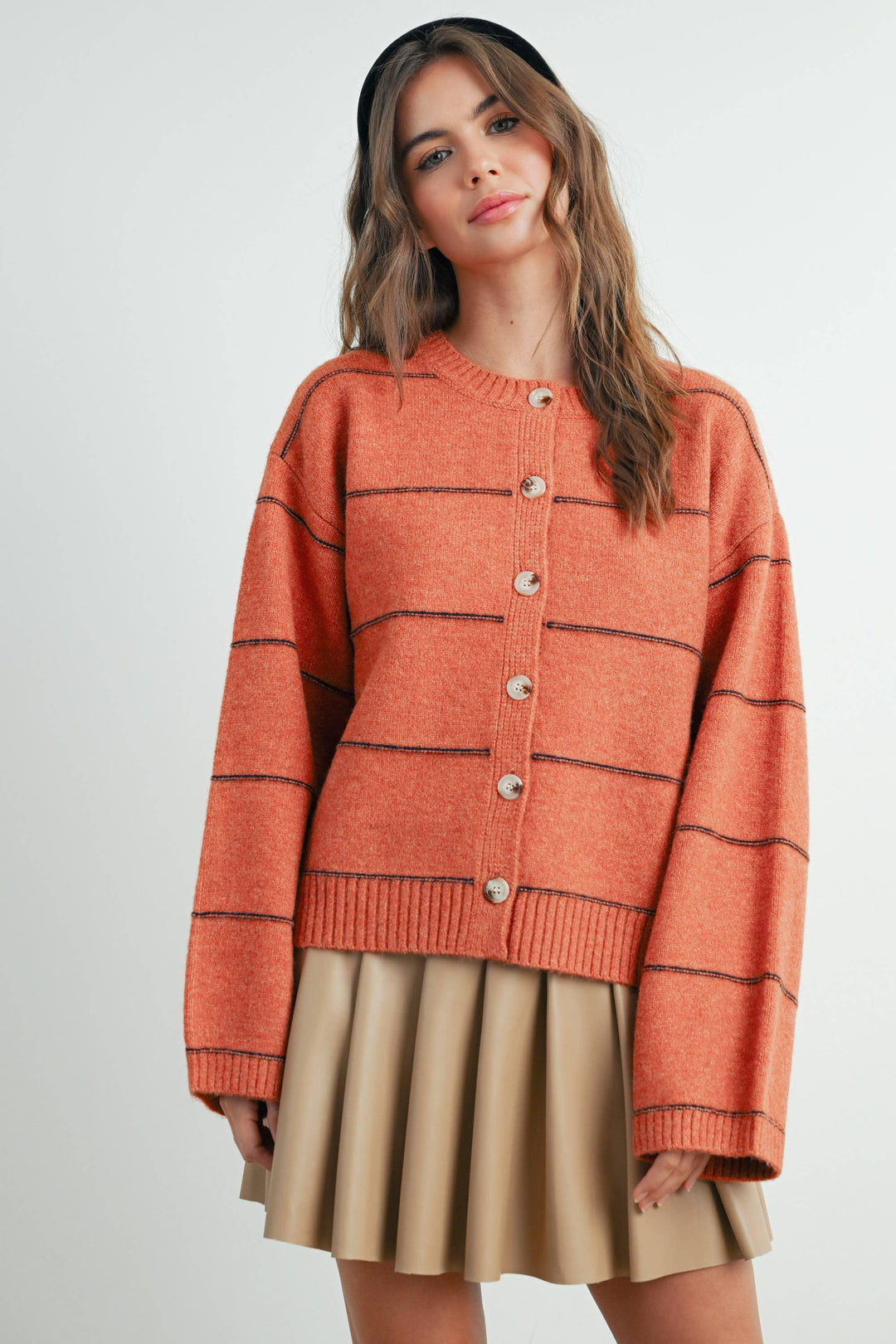 Rust orange button-up cardigan with black stripes, paired with beige pleated skirt and black headband