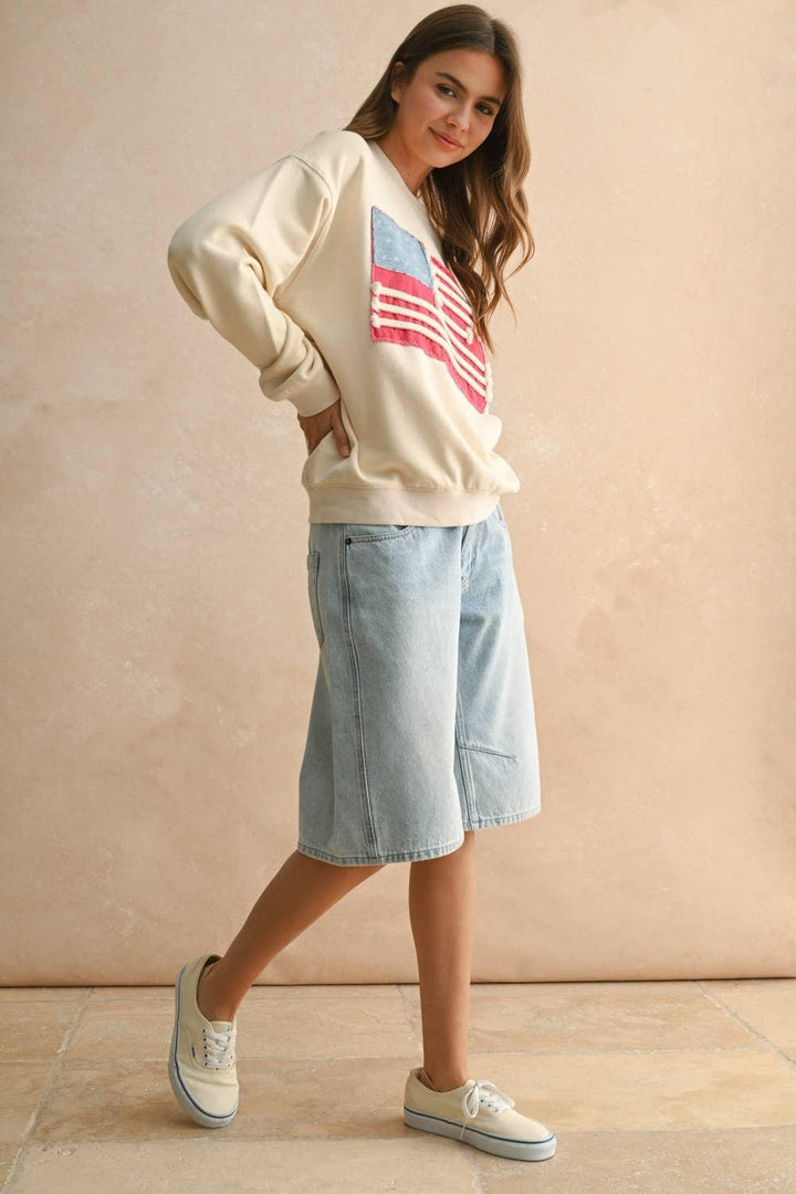 Woman wearing a MIOU MUSE cream sweatshirt with a red, white, and blue flag patch and denim skirt.