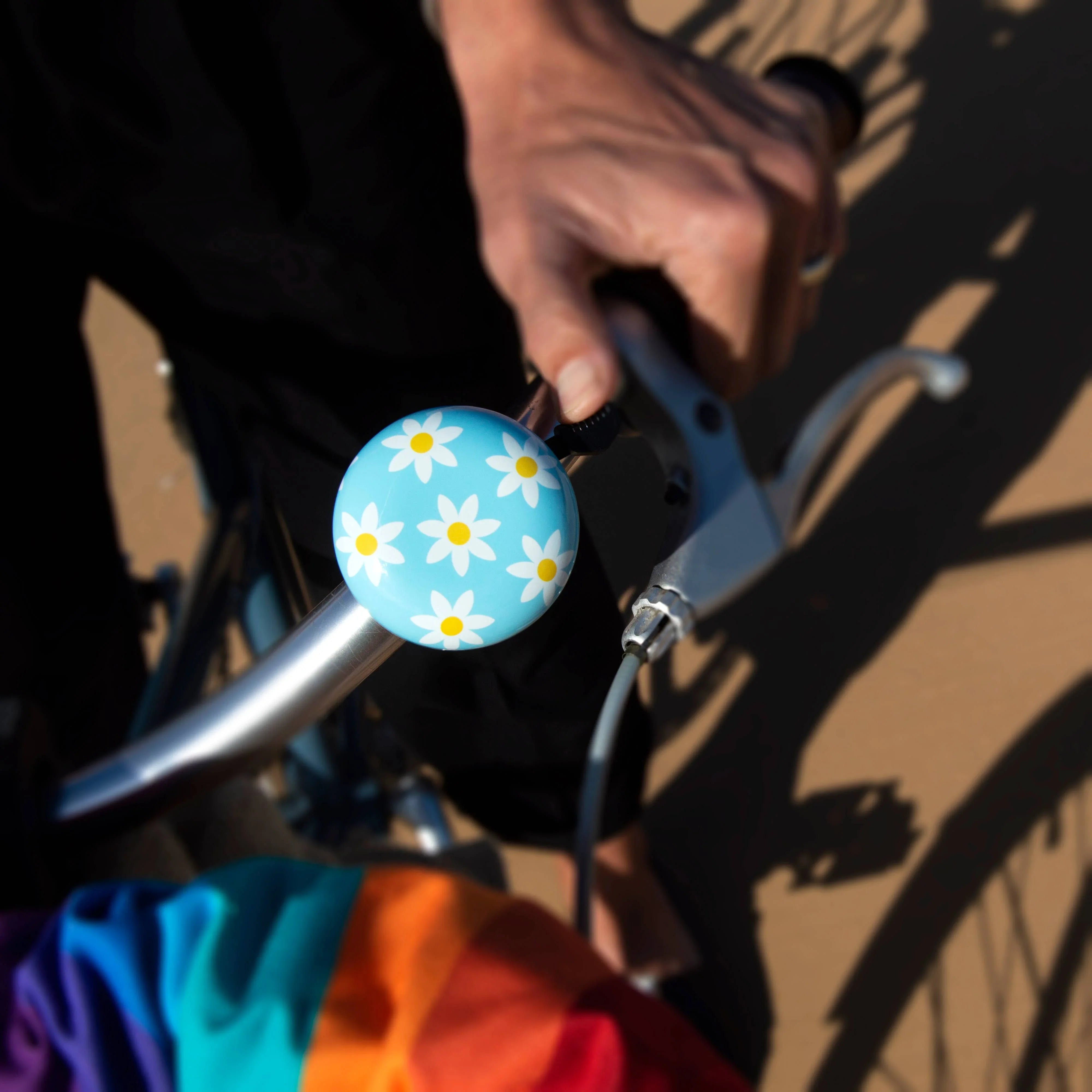 Blue daisy-patterned bike bell on handlebars, hand gripping brake, rainbow bag, coastal ride gear