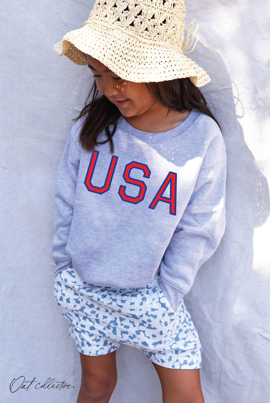Child in USA graphic sweatshirt, patterned shorts, and crocheted sun hat for coastal adventures