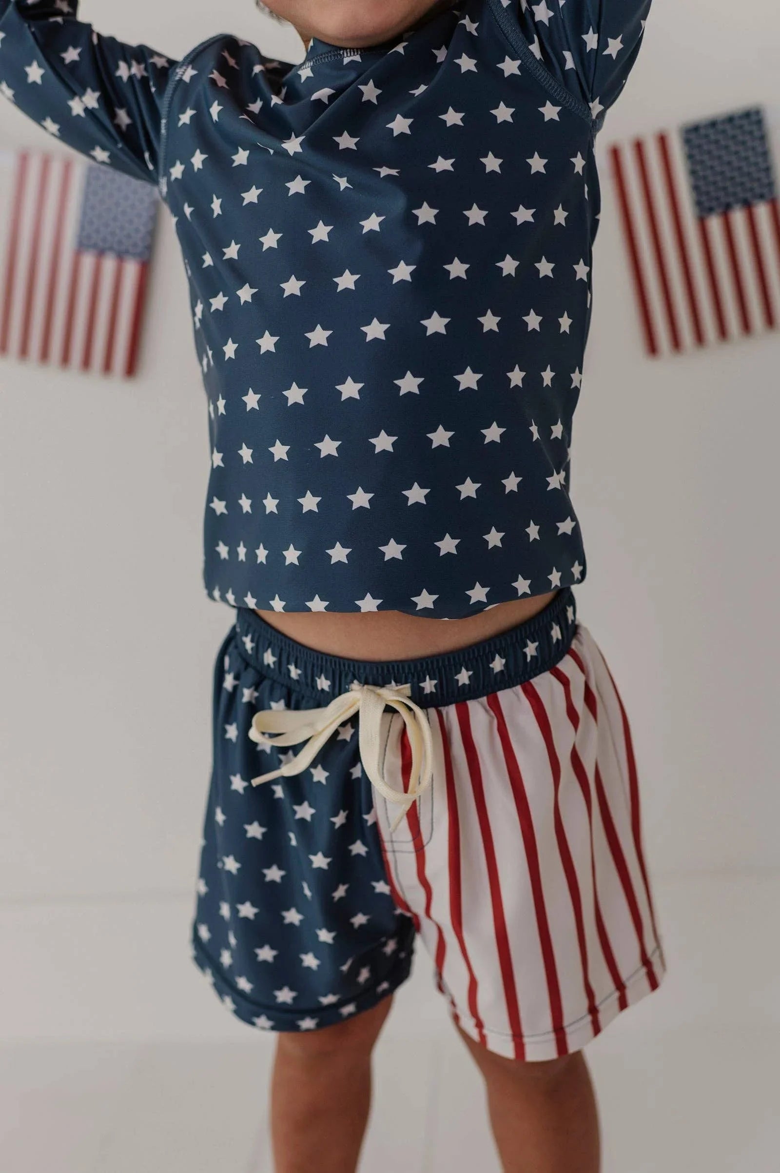 Child in navy star rash guard and red-white striped swim trunks, patriotic beachwear for summer adventures