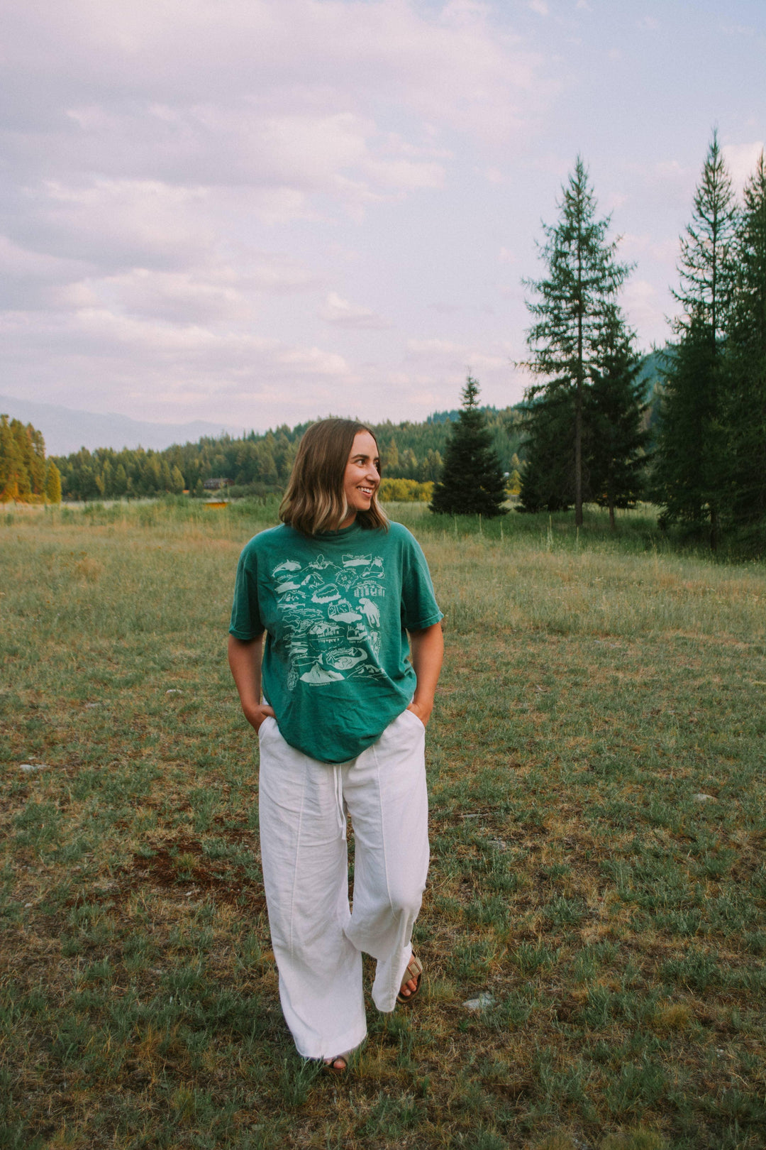Woman in green PNW map tee &amp; white pants, smiling outdoors in grassy field with trees