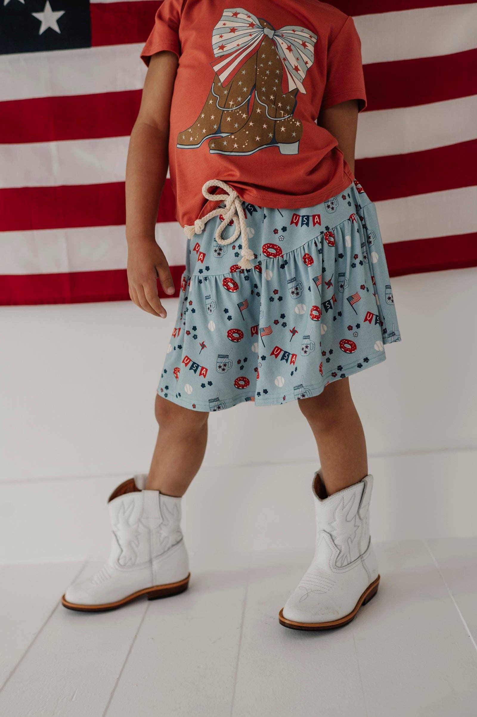 Child in red cowboy boot tee, USA print skirt, white boots, American flag background