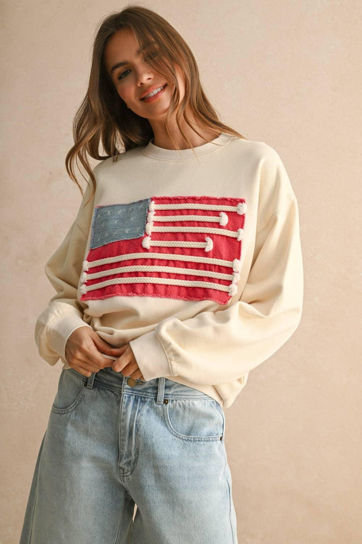 Woman wearing a cream MIOU MUSE sweatshirt with a red and blue flag patch and light wash jeans.