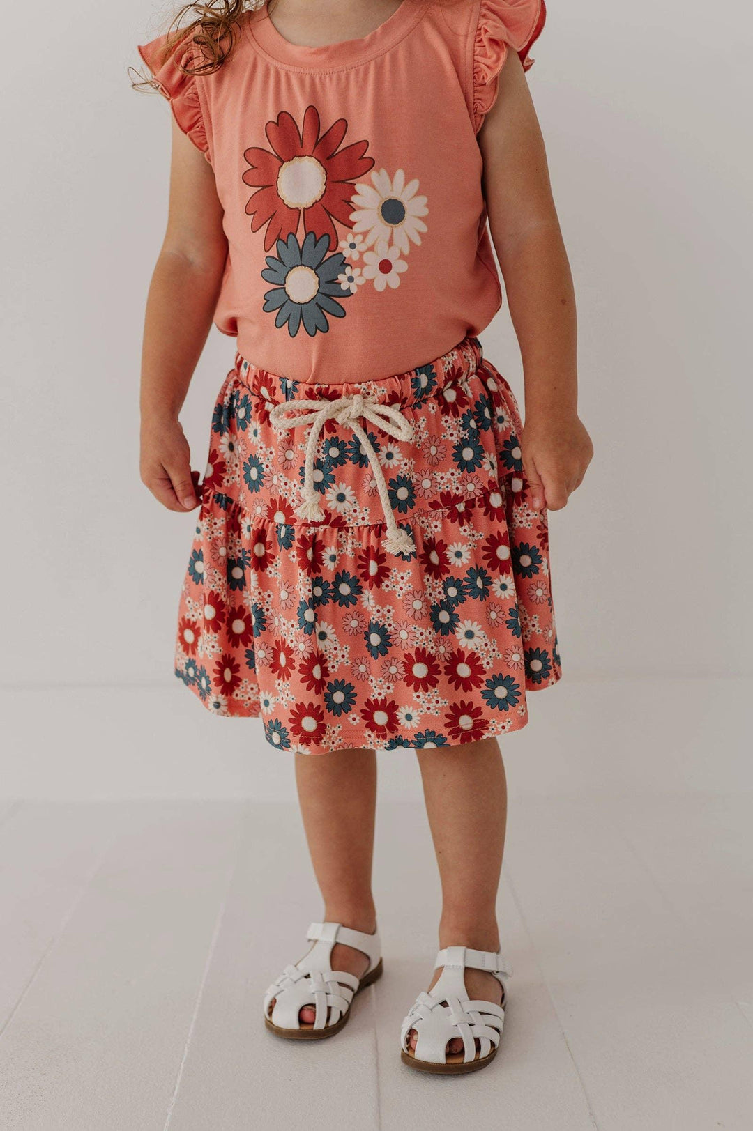 Toddler in coral floral top and matching skirt with white sandals, perfect for beach days and outdoor adventures.