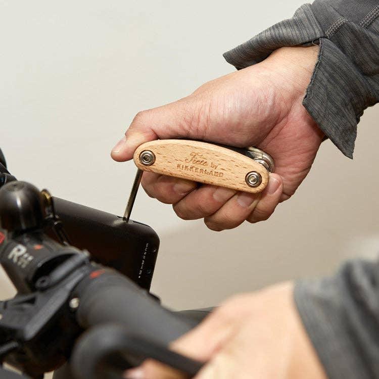 Hand using wooden multi-tool to adjust bike handlebar mount, ideal for outdoor adventures and trail rides.