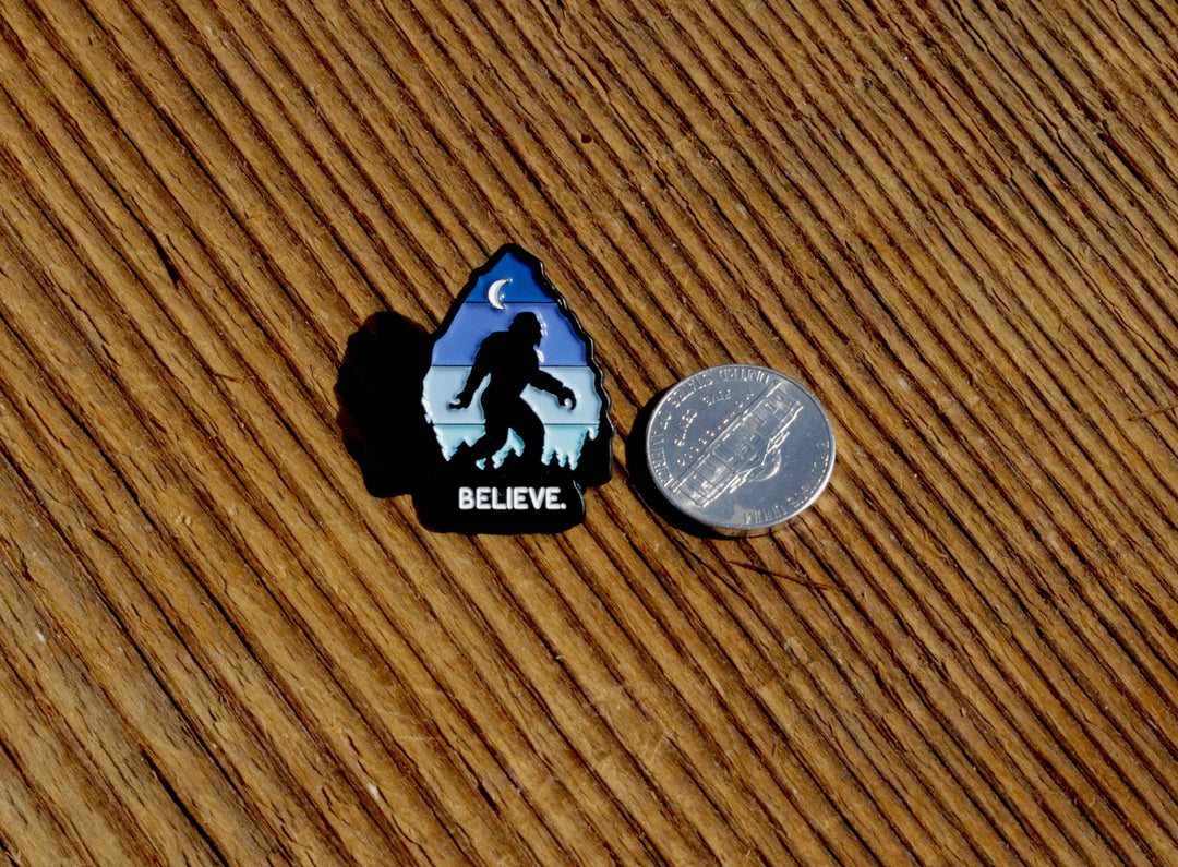 Believe Bigfoot enamel pin with moon and mountains, shown beside a quarter for scale on wood.