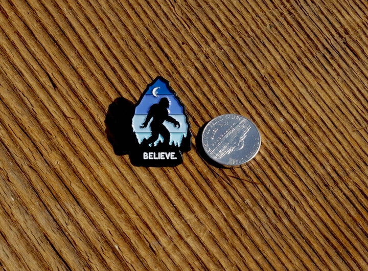 Believe Bigfoot enamel pin with moon and mountains, shown beside a quarter for scale on wood.