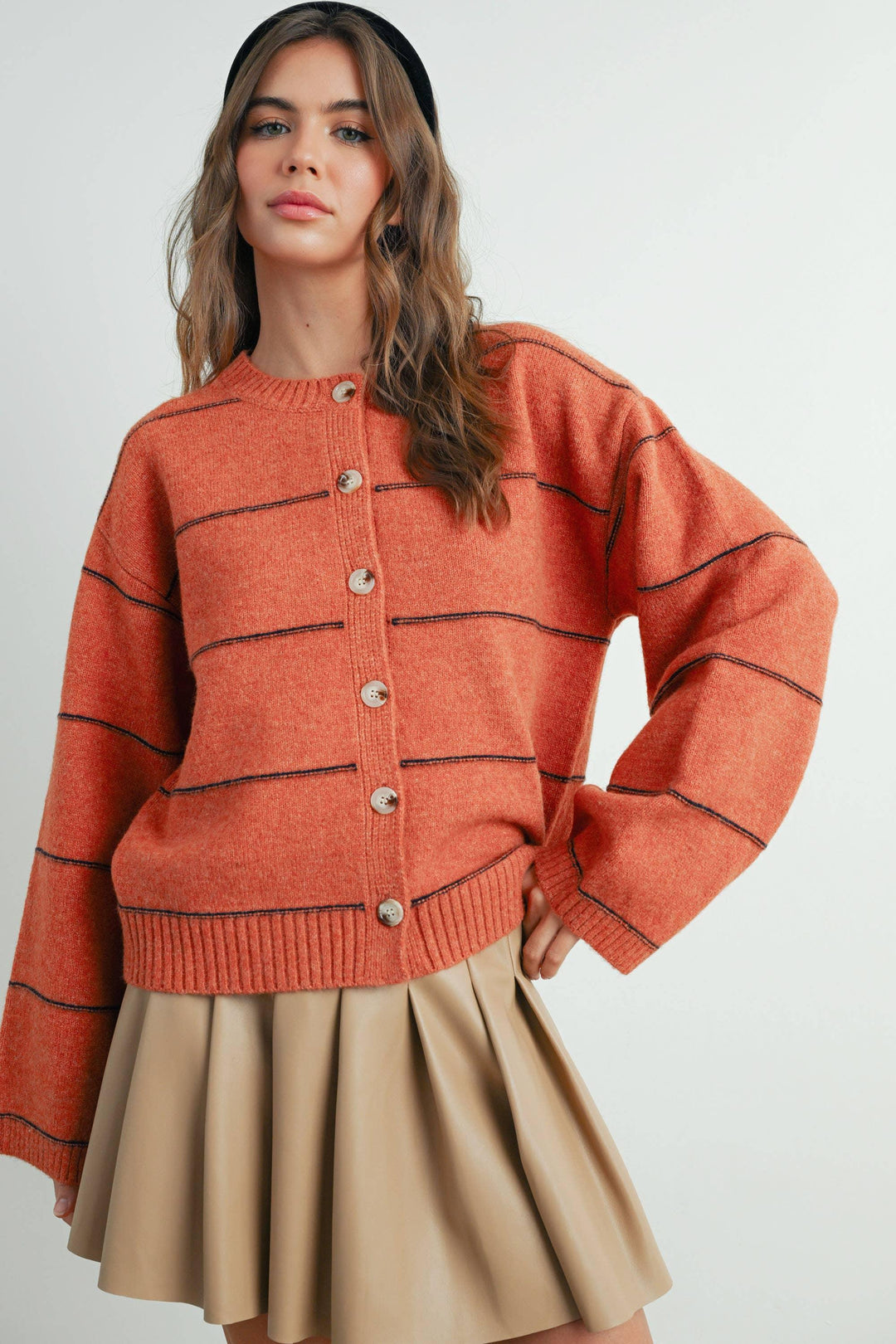 Rust orange button-up cardigan with black stripes, paired with beige pleated skirt and black headband