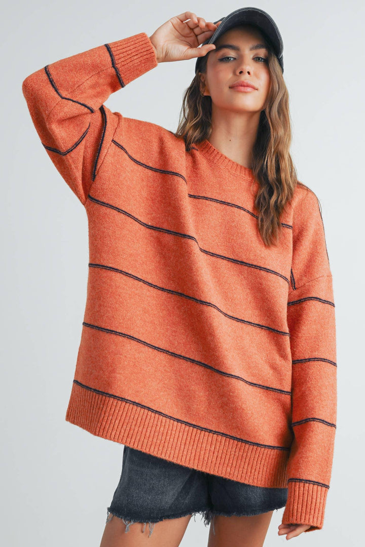 Orange striped knit sweater with black trim, paired with denim shorts and a cap for casual fall style