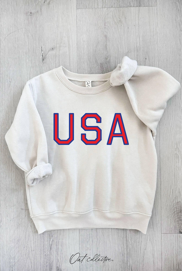 Cream USA graphic sweatshirt with red and blue lettering, laid on wood floor