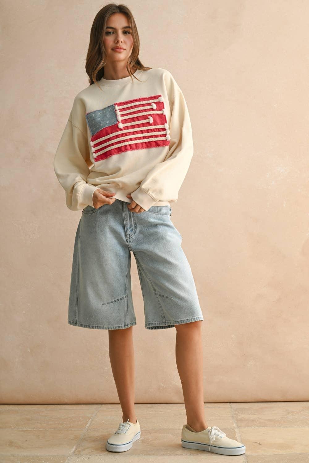Woman wearing a cream MIOU MUSE sweatshirt with a red and blue flag patch and denim shorts.