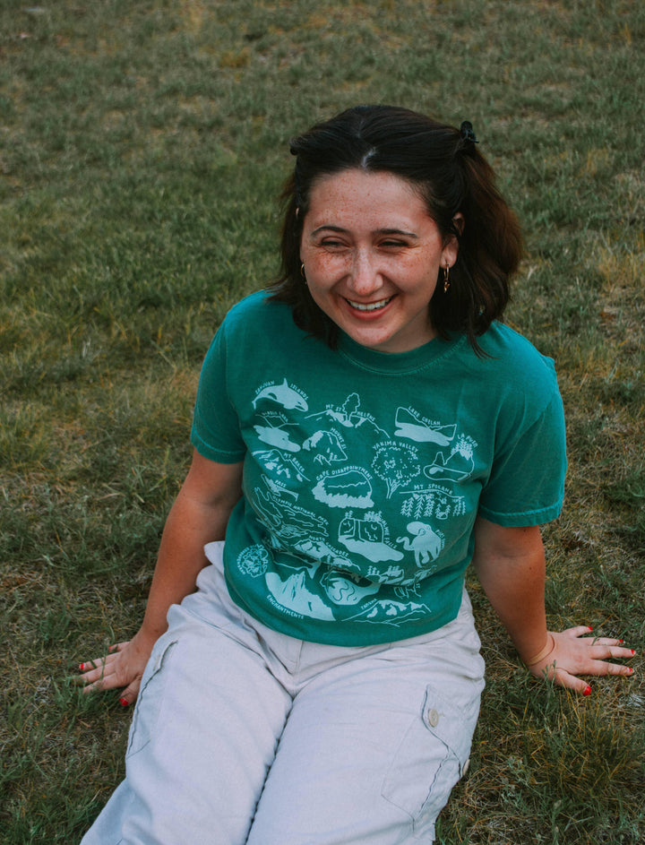 Woman smiling in green PNW map tee, sitting on grass, outdoor adventure style