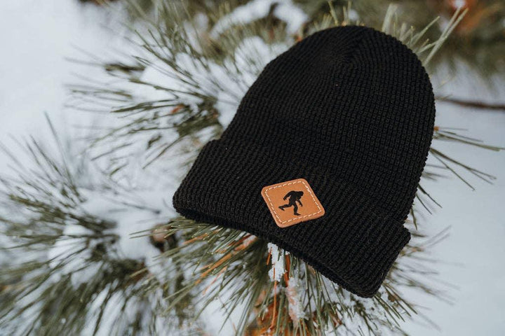 Black knit beanie with leather patch featuring Sasquatch logo, nestled in snowy pine branches