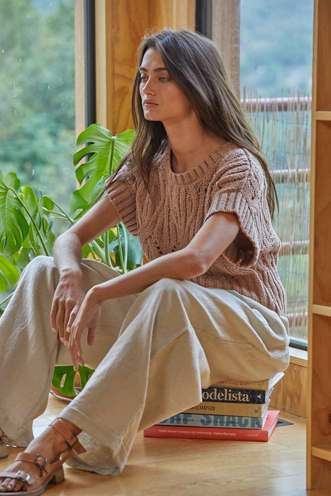 Woman in cozy knit sweater and wide-leg pants, sitting by window with green plants, relaxed home style
