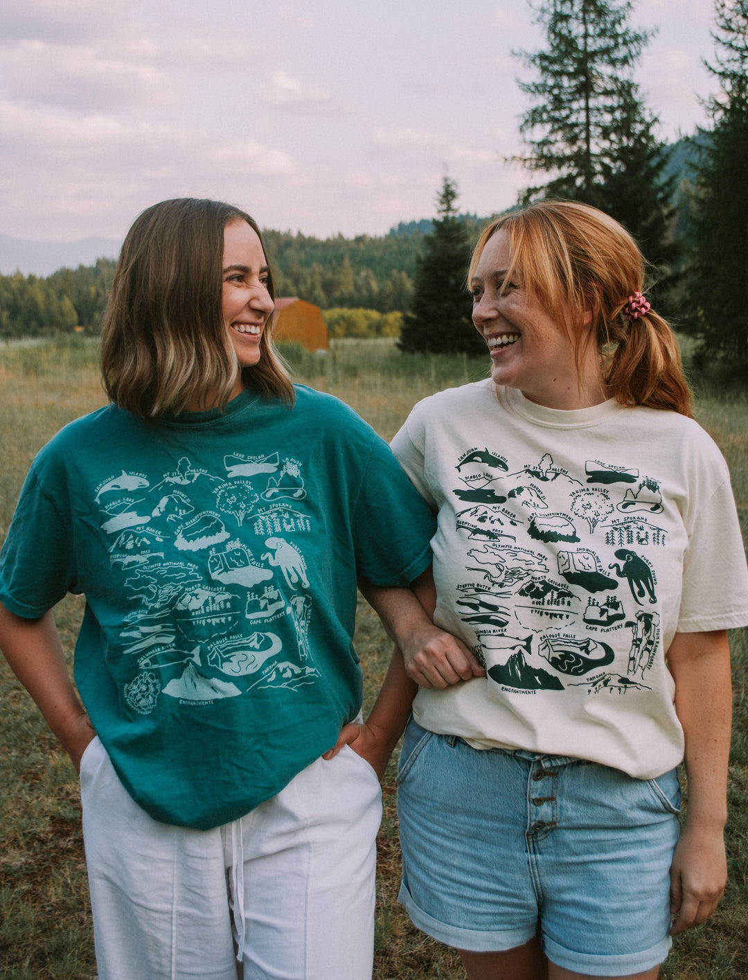 Two women in PNW adventure graphic tees smiling outdoors near forest, perfect for coastal & trail weekends.
