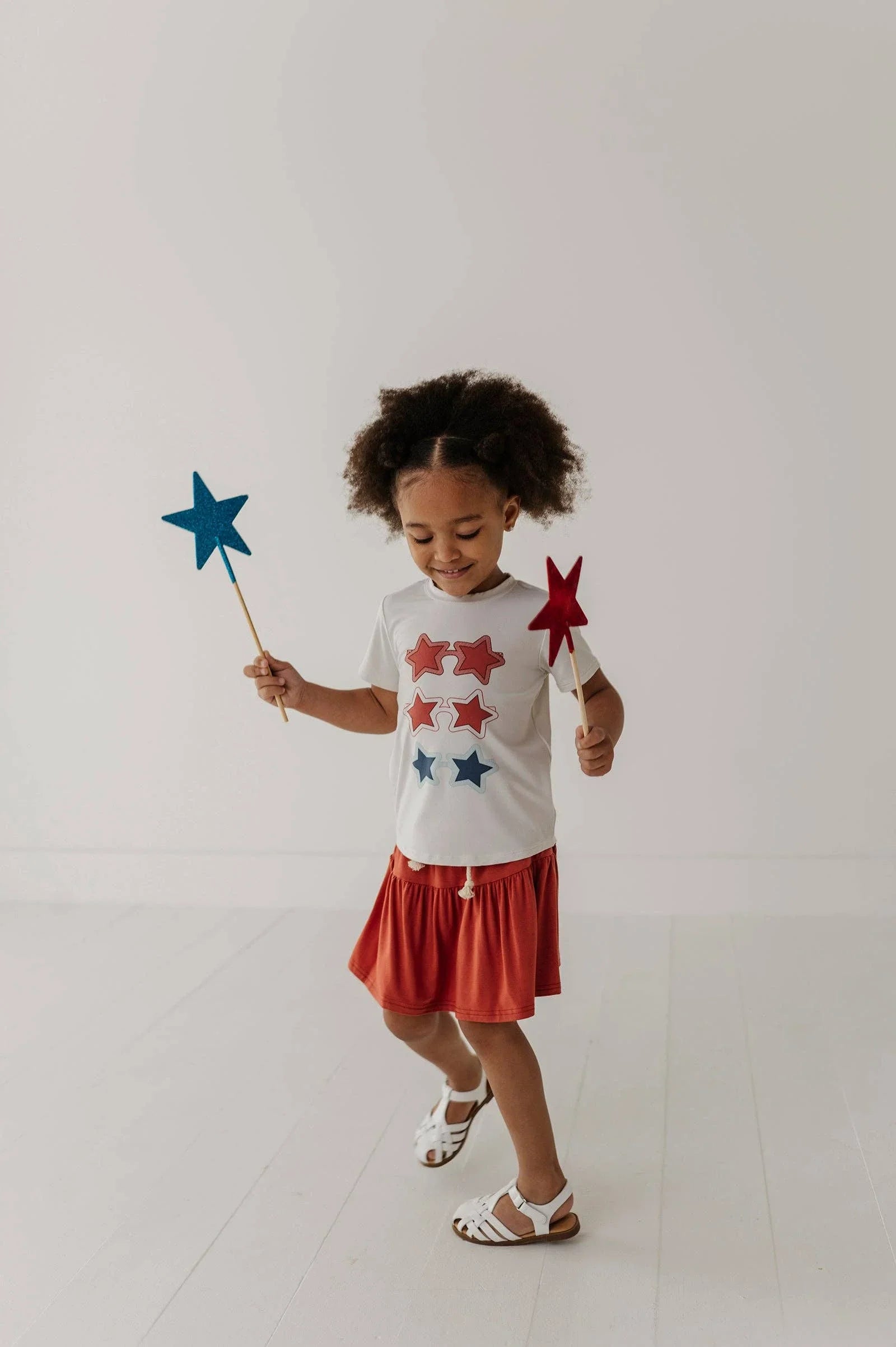 Toddler in star-print tee & red skirt holding blue and red star wands, playful summer outfit