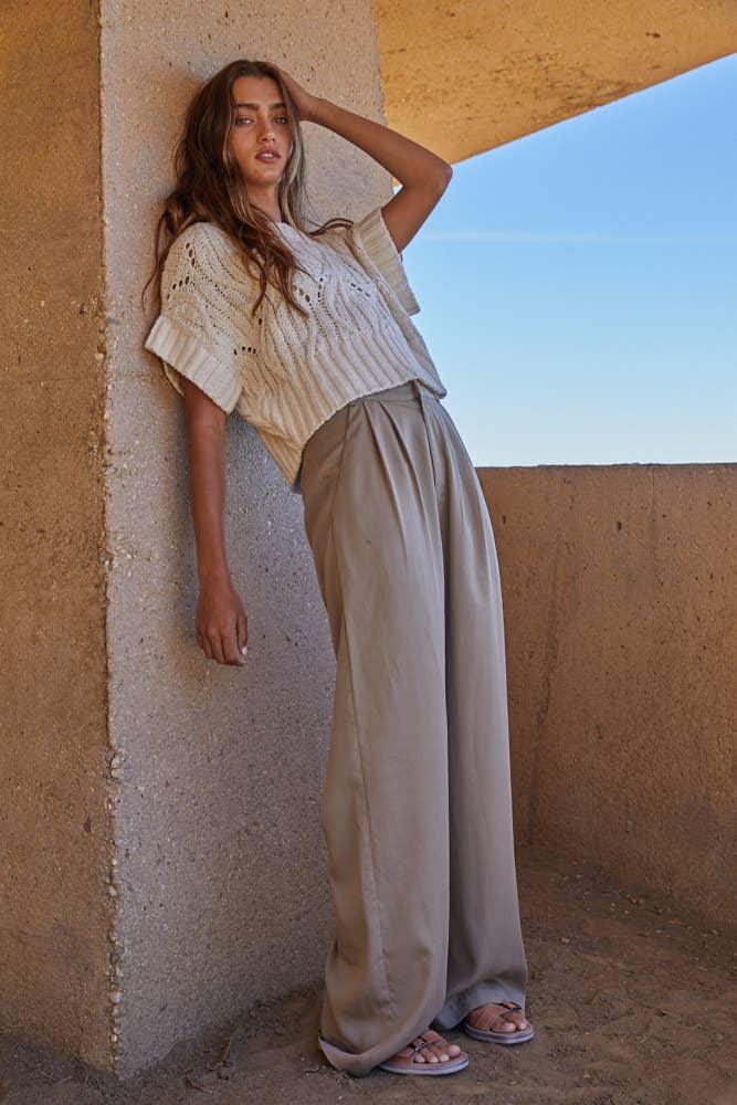 Woman in beige knit top and wide-leg pants leaning against concrete wall outdoors