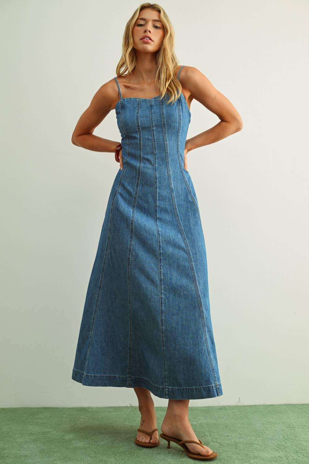 Woman in blue denim midi dress with thin straps and side pockets, standing on green floor