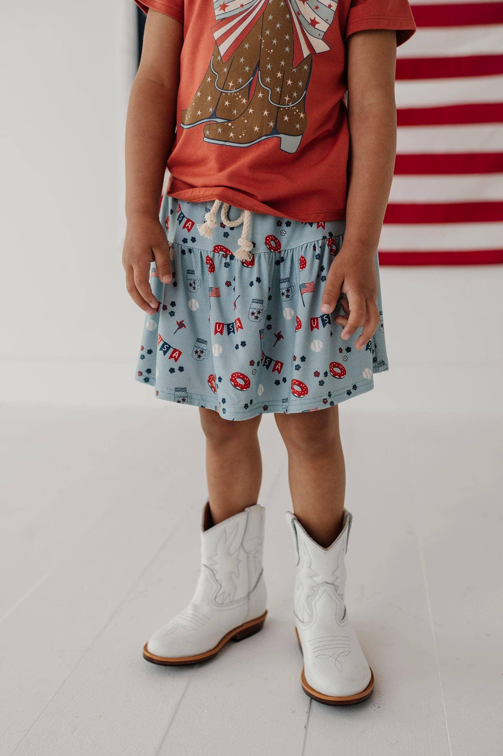 Child in red USA graphic tee, light blue patriotic skirt, and white cowboy boots, standing before American flag.