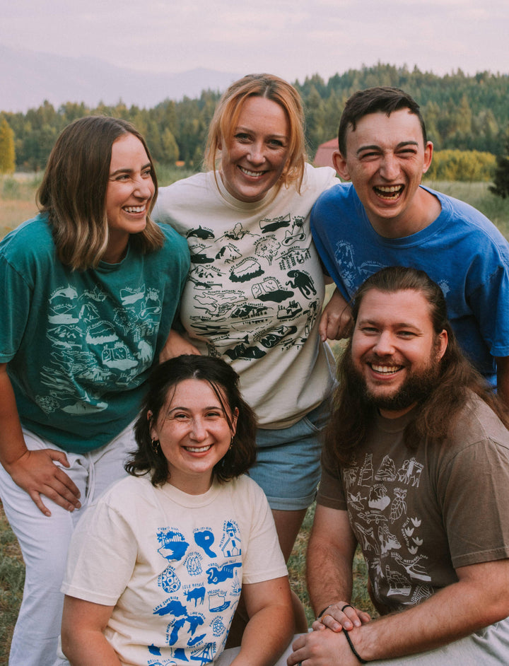 Friends in PNW adventure t-shirts smiling outdoors near forest, perfect for trail hikes and coastal weekends.