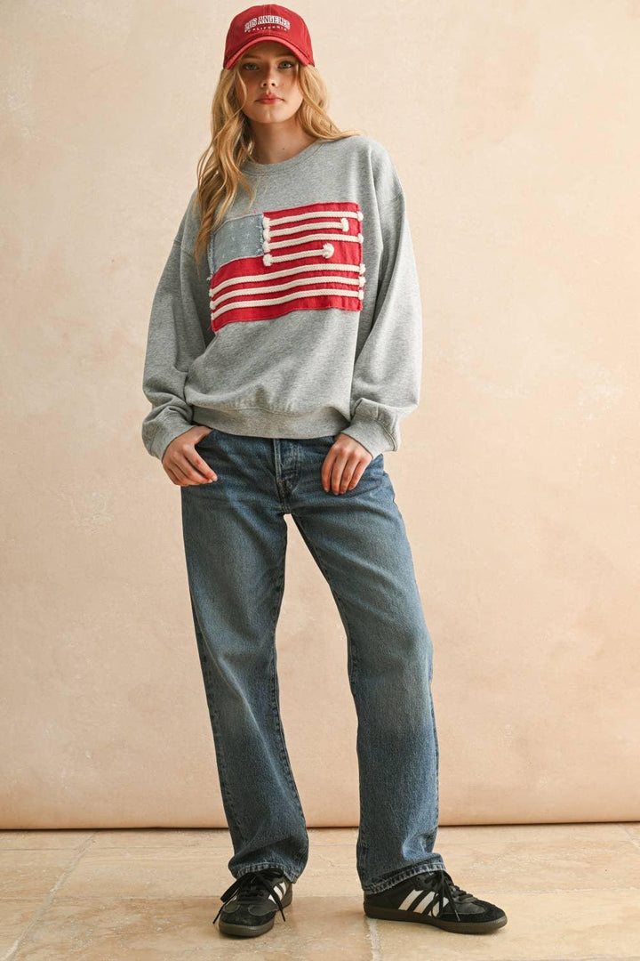 Woman wearing a MIOU MUSE grey sweatshirt with a red and white flag patch and blue jeans.
