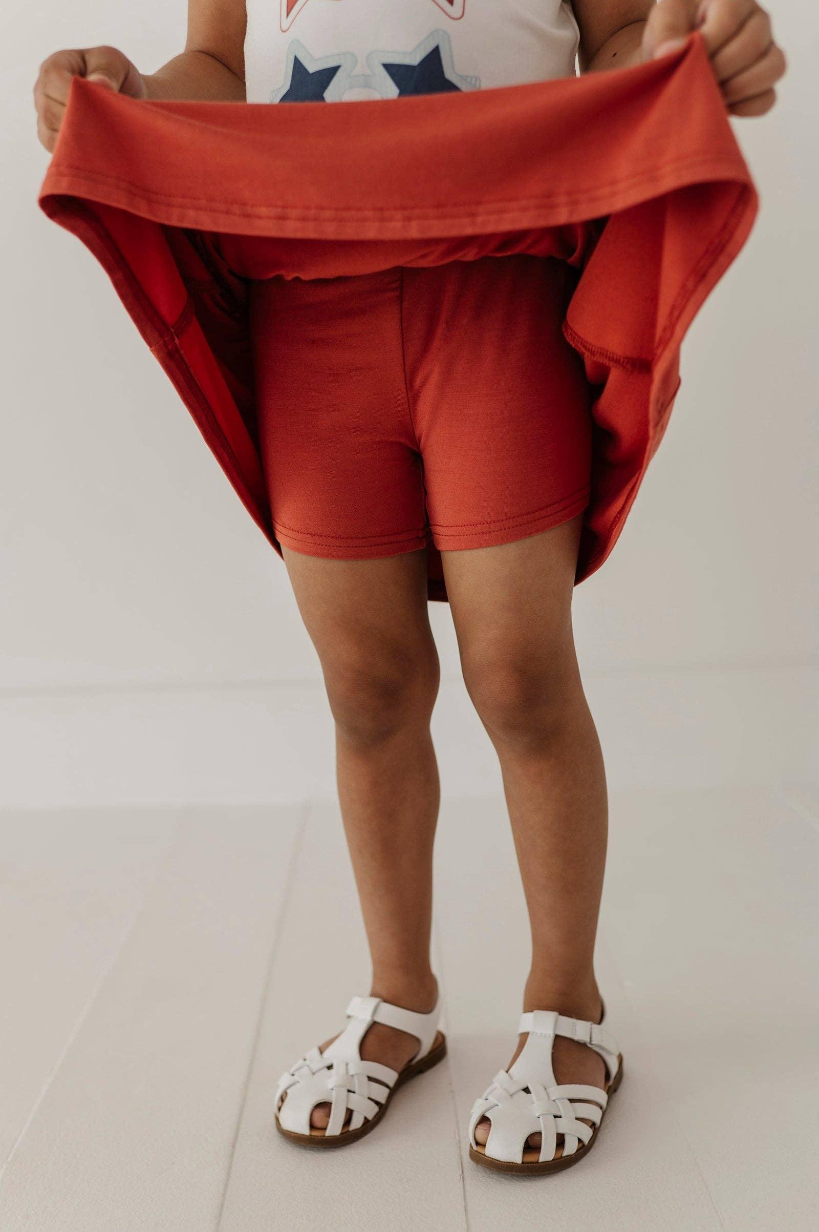 Child in rust red shorts and white sandals, holding up matching top for coastal PNW adventures