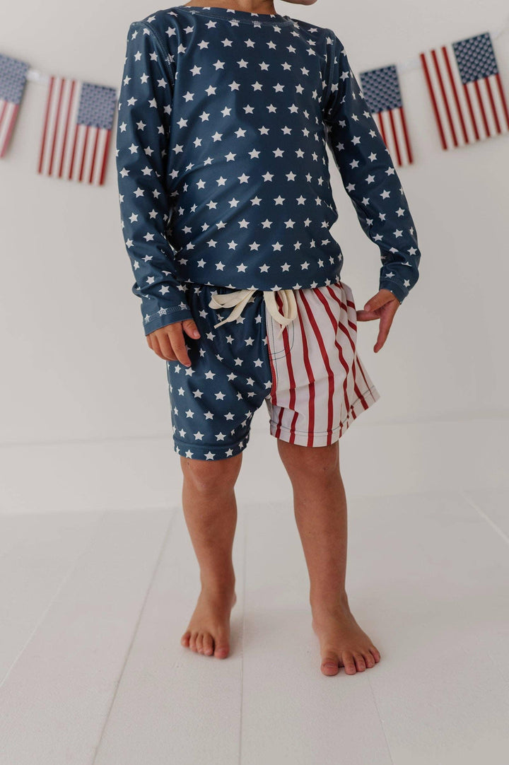 Kid in stars and stripes swim set with American flag bunting, perfect for beach days and coastal adventures.