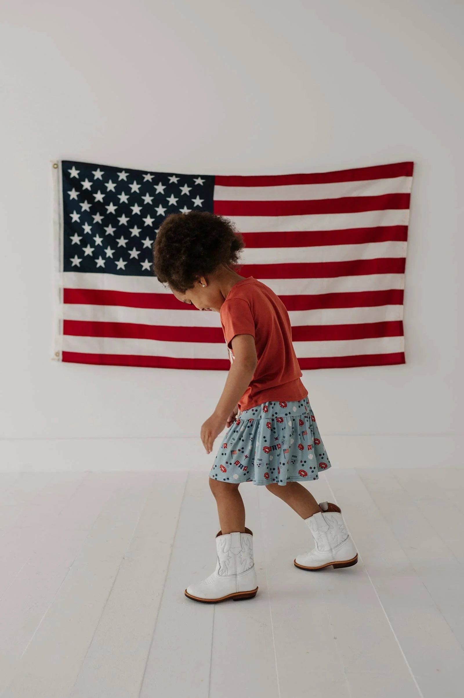 Child in red tee, patterned skirt, white cowboy boots, walking past American flag