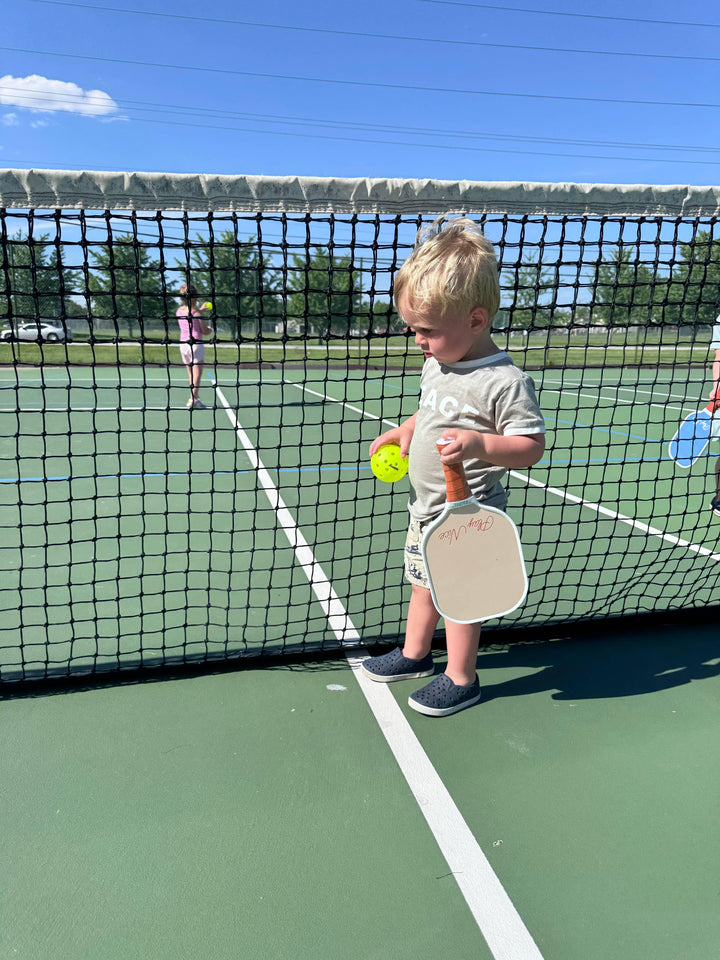 Toddler holding pickleball paddle and ball on outdoor court, ready for family fun under sunny skies