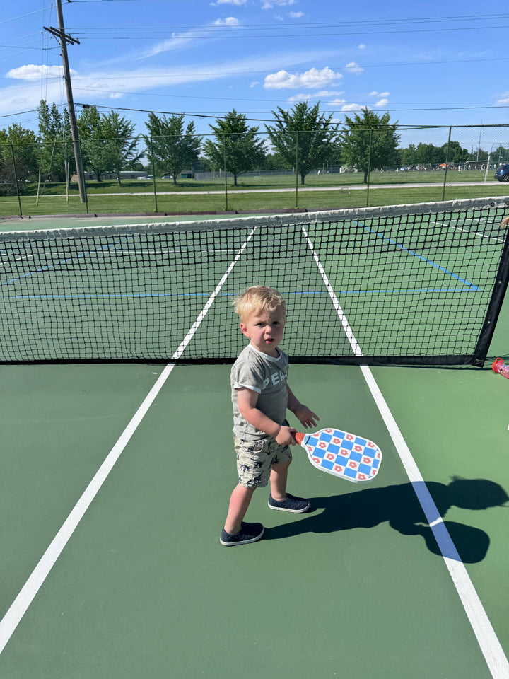 Toddler holding colorful pickleball paddle on outdoor court under sunny sky